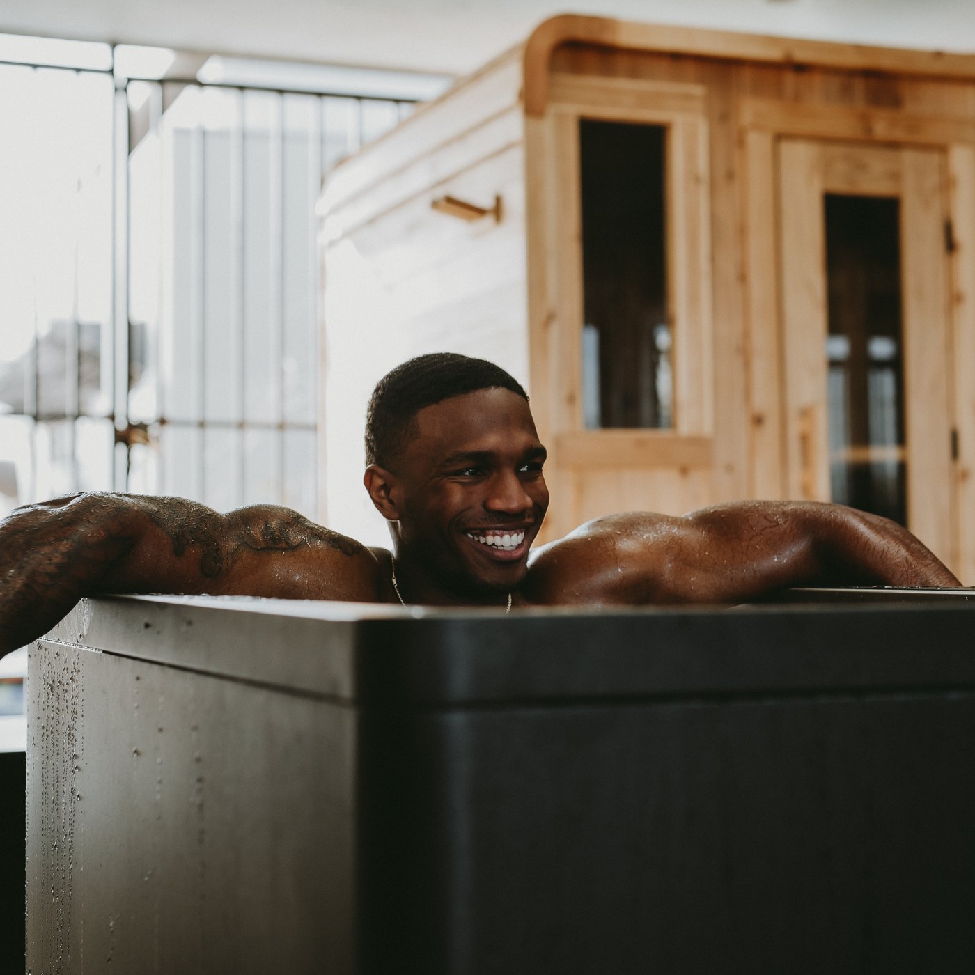 A WLF member resting in the cold plunge with his arms over the sides, smiling, the cedar sauna behind him.
